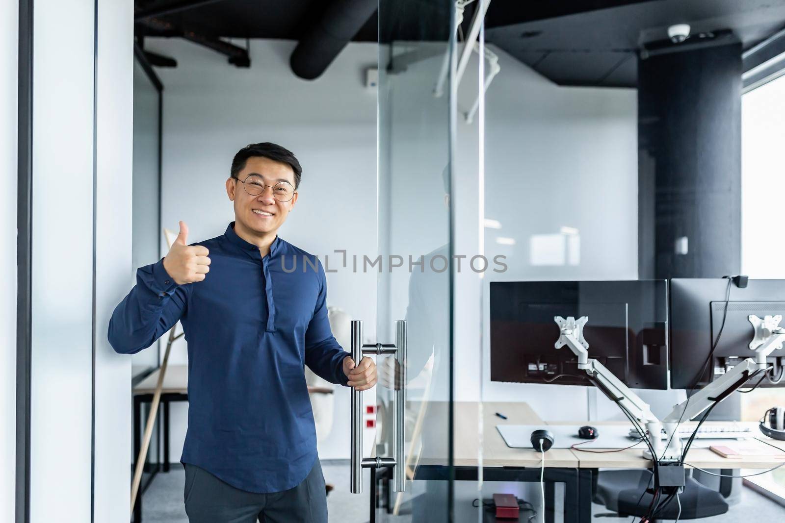 Portrait of Asian designer engineer, man smiling and looking at camera holding thumb up affirmative, businessman working with several computers inside twisted office building.