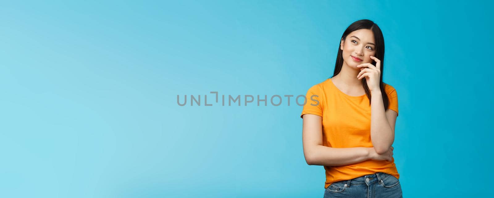 Joyful confident asian girl with dark haircut tilt head, dreaming, imaging interesting plan, smiling cunning, have idea, thinking and making choice, look up thoughtful, stand blue background.