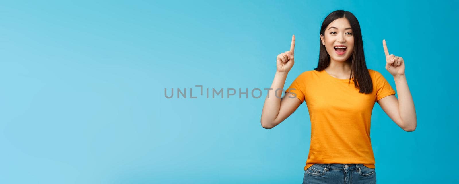 Amused asian girl introduce new product sharing promo with you, raise hands pointing fingers up smiling broadly, look excited and upbeat, enthusiastic advertising, stand blue background.