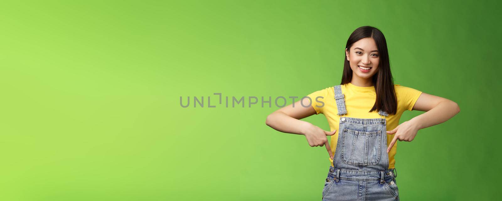 friendly outgoing cute asian young woman show fingers down, introduce new promo app, smiling joyfully sharing link, recommend product using brand, grinning toothy, green background. Copy space