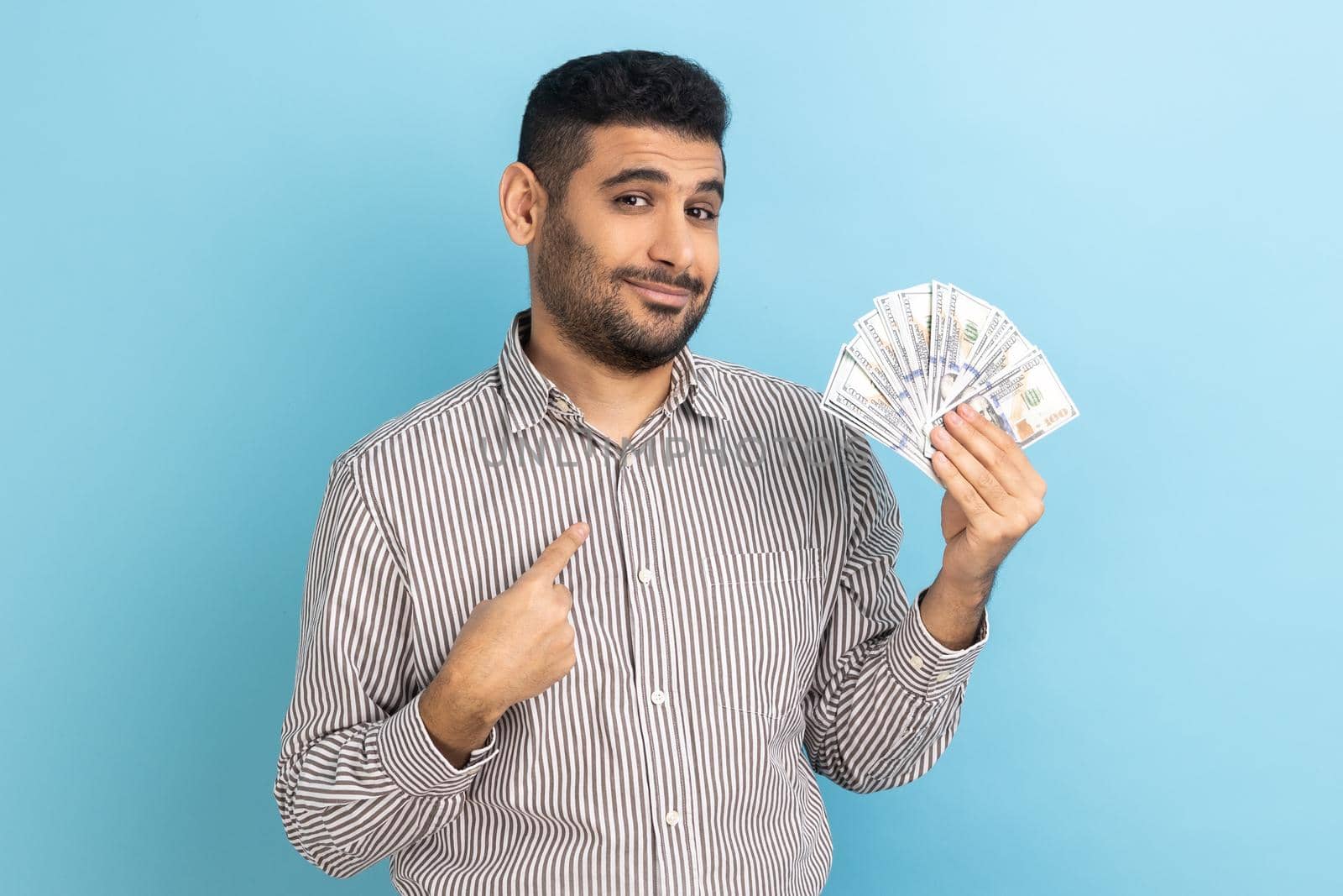 Portrait of young adult businessman with beard holding dollar bills and pointing at himself with pride, looking at camera, wearing striped shirt. Indoor studio shot isolated on blue background.