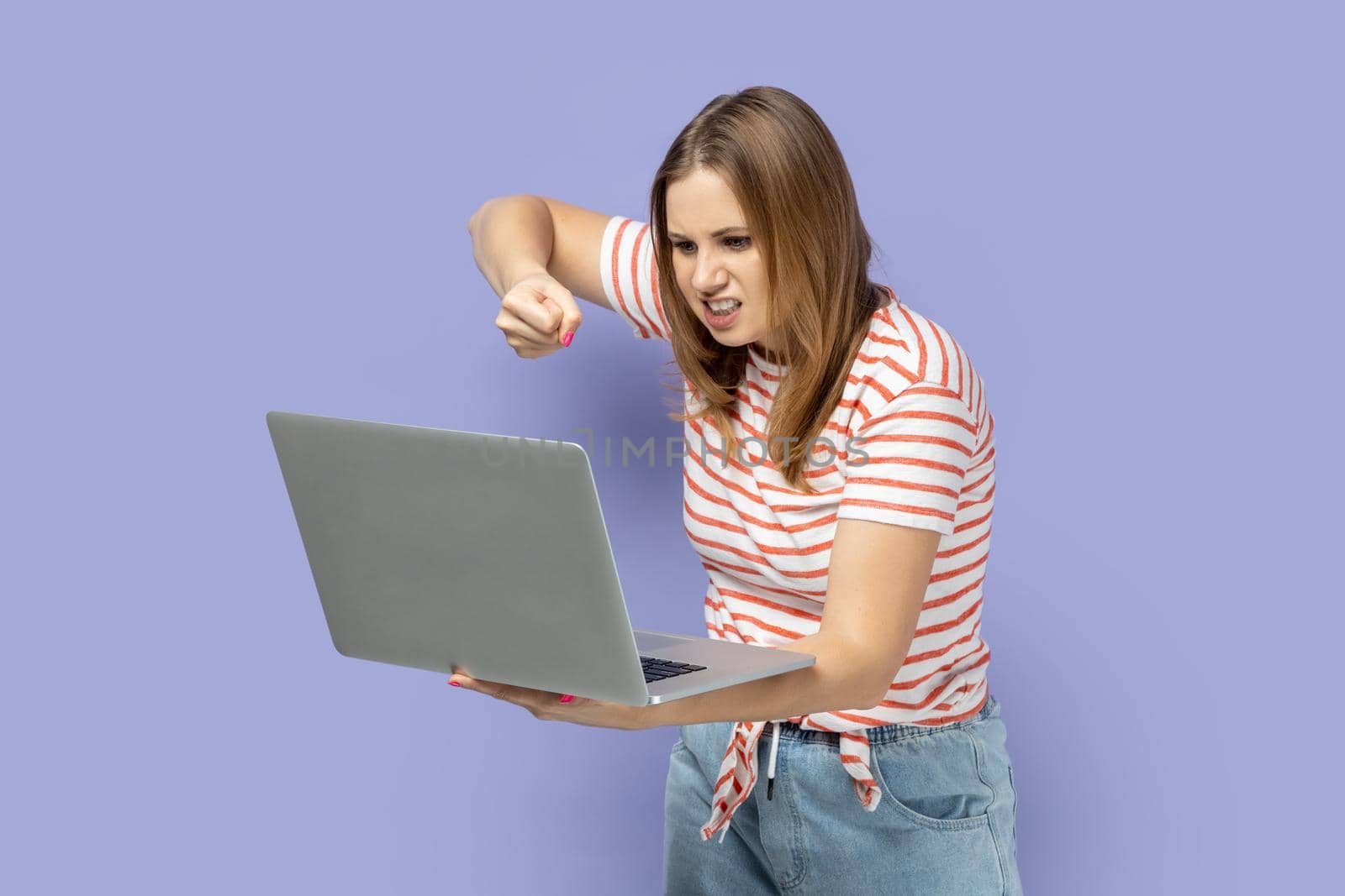 Portrait of angry aggressive blond woman wearing striped T-shirt working on laptop, having online conversation, boxing to notebook display. Indoor studio shot isolated on purple background.