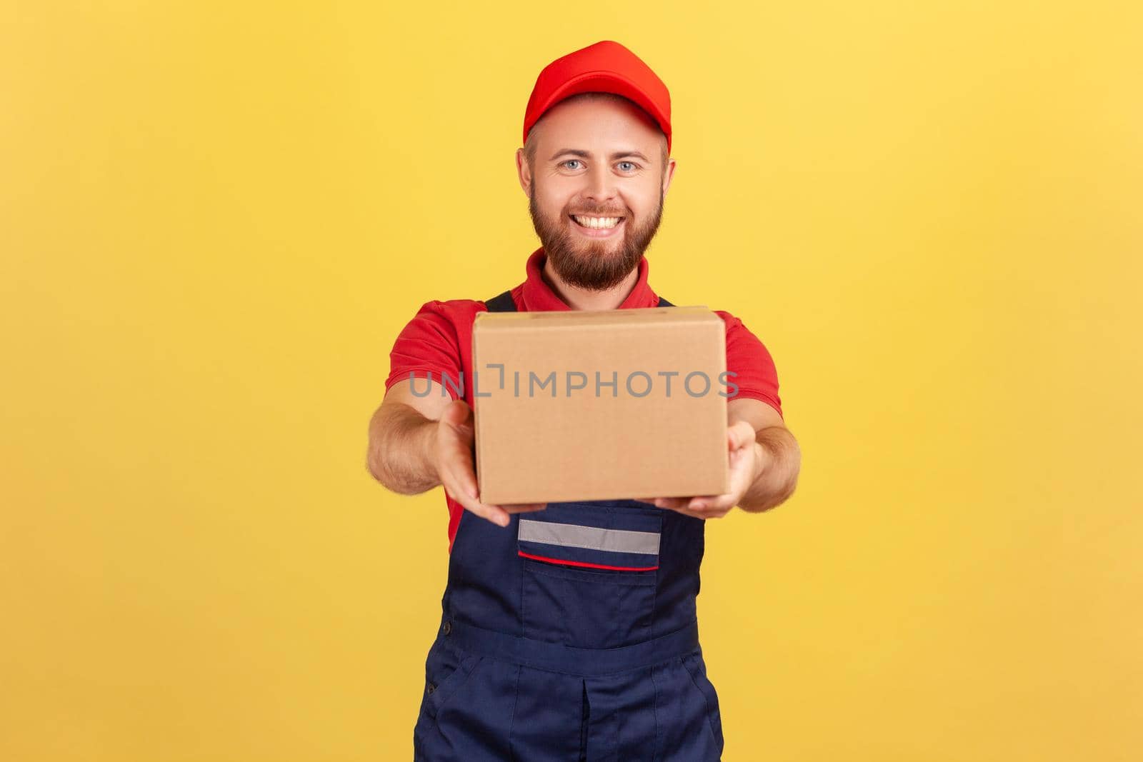 Portrait of smiling courier man holding cardboard parcel, delivering order door-to-door, shipment and cargo transportation service. Indoor studio shot isolated on yellow background.