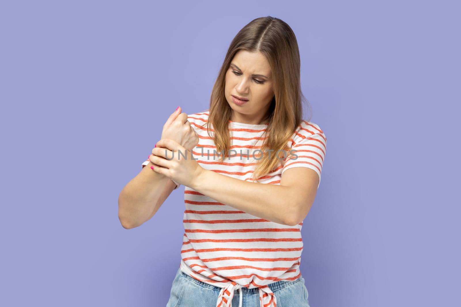 Unhappy sick blond woman wearing striped T-shirt touching painful hand, suffering trauma, sprain wrist, feeling ache of carpal tunnel syndrome. Indoor studio shot isolated on purple background.