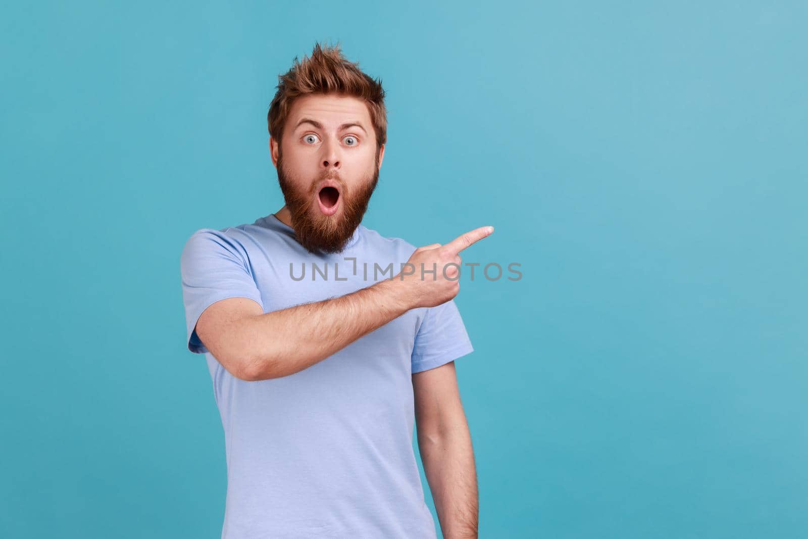 Portrait of surprised shocked bearded man pointing aside, showing blank copy space for idea presentation, commercial text, keeps mouth open. Indoor studio shot isolated on blue background.