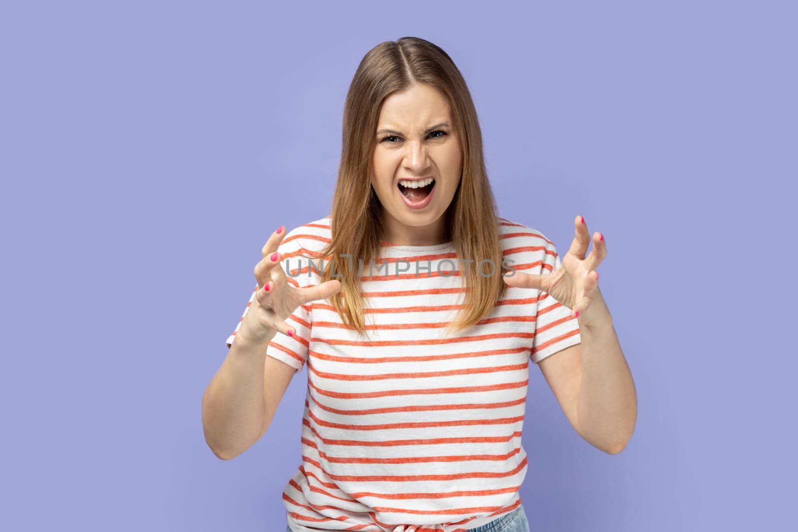 Portrait of angry young adult blond woman wearing striped T-shirt expressing aggressive emotions, raised arms, screaming with hate and anger. Indoor studio shot isolated on purple background.