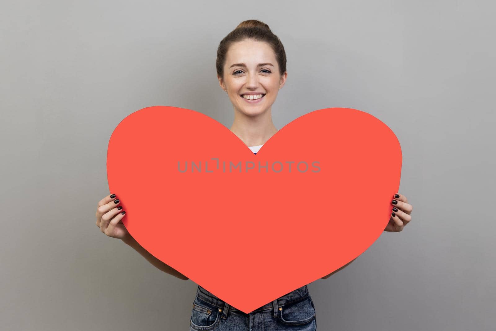 Portrait of happy woman smiling at camera and holding big red heart, symbol of love and affection, expressing i love you and romantic feelings. Indoor studio shot isolated on gray background.