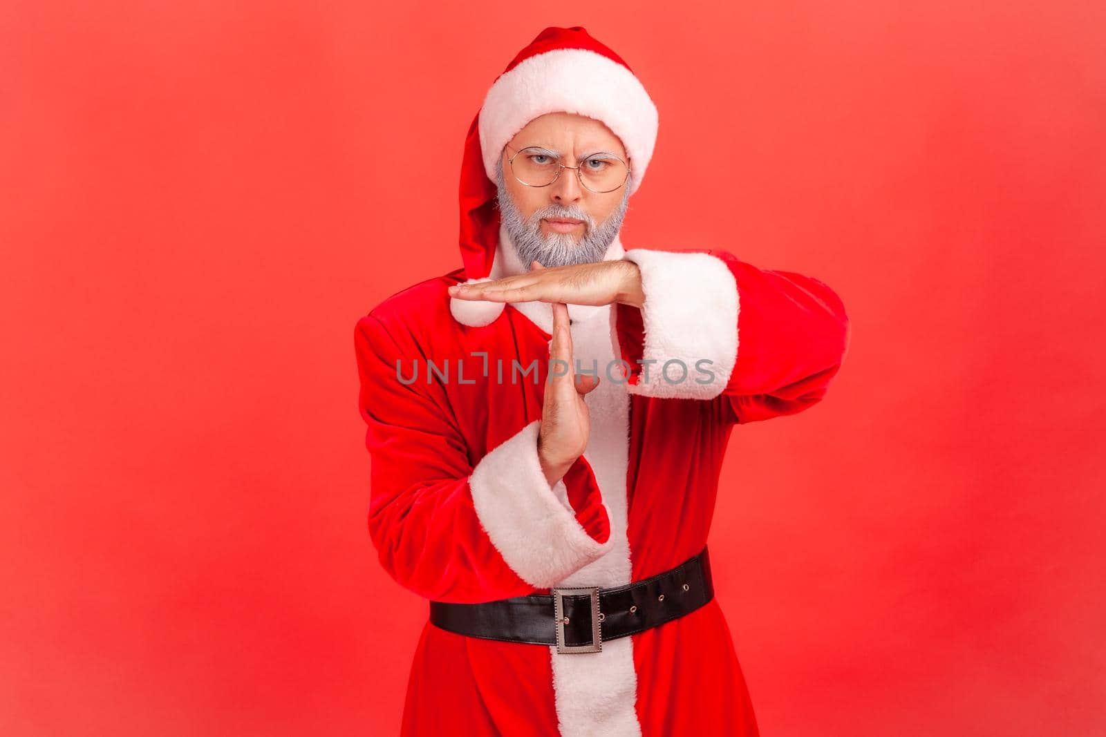 Please, I need break. Elderly man with gray beard wearing santa claus costume showing time out gesture, stop or limit hand sign, warning of deadline. Indoor studio shot isolated on red background.