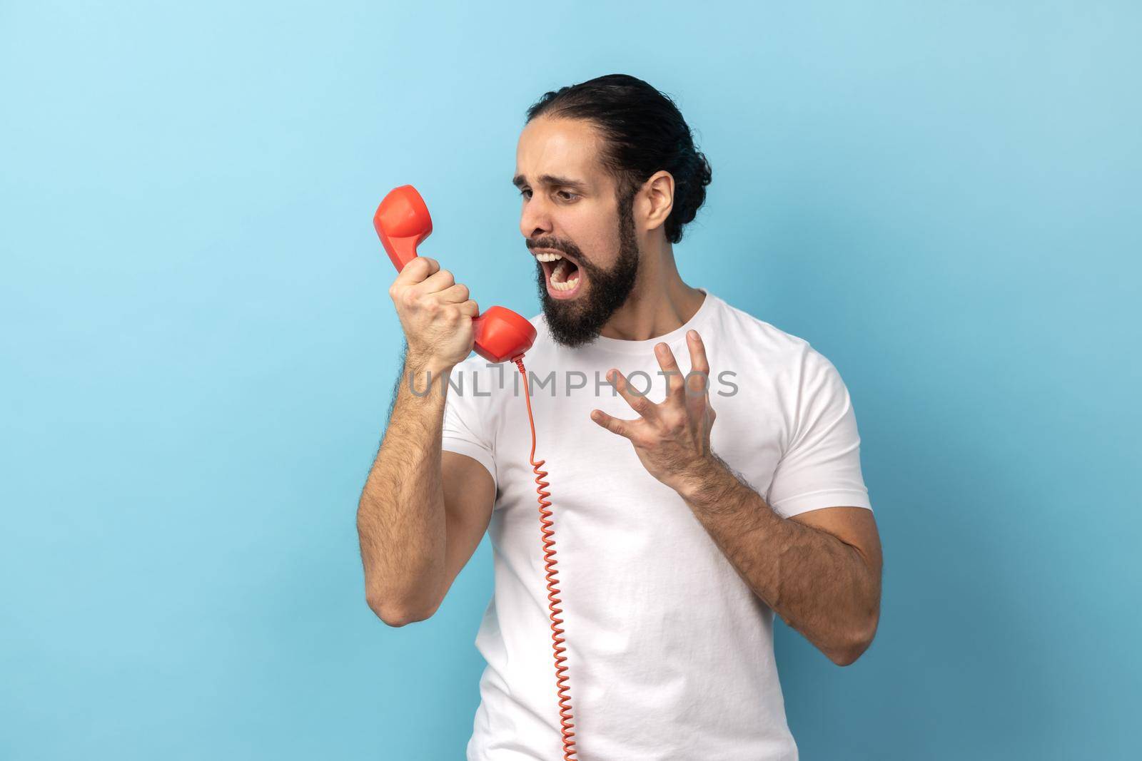 Portrait of angry man with beard wearing white T-shirt screaming and yelling talking retro landline phone, complaining on connection quality. Indoor studio shot isolated on blue background.