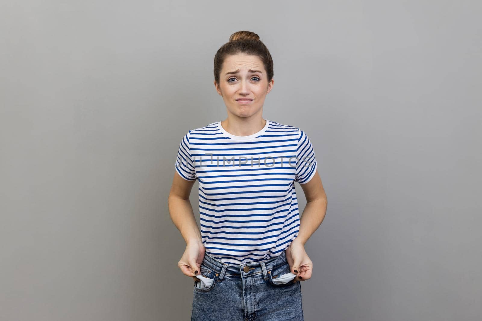 Portrait of upset poor attractive woman wearing striped T-shirt turning out empty pockets, showing no money gesture, worried about debts. Indoor studio shot isolated on gray background.