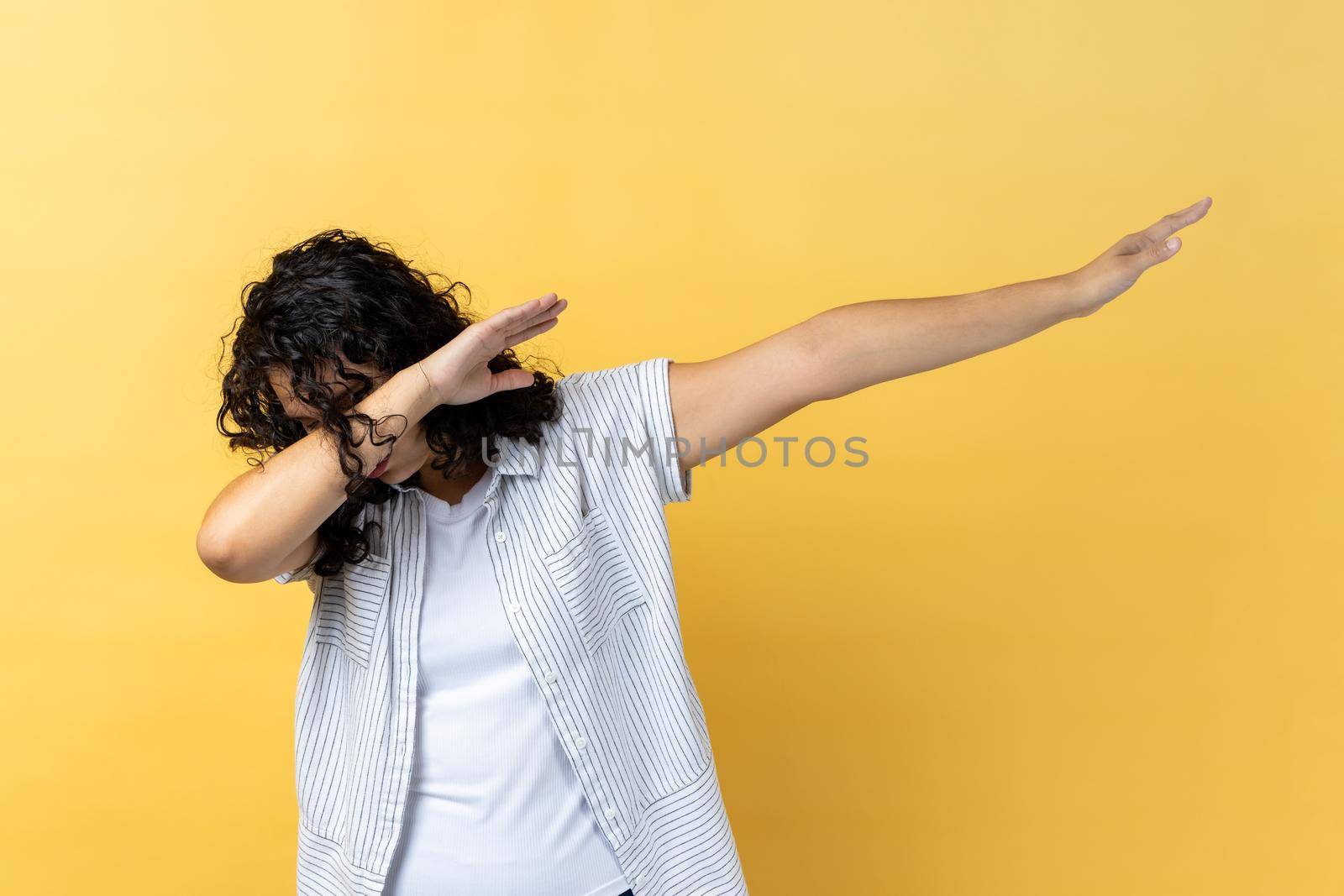 Portrait of young woman with dark wavy hair, hiding face with dab dance move, performing internet meme of success, dabbing trends. Indoor studio shot isolated on yellow background.