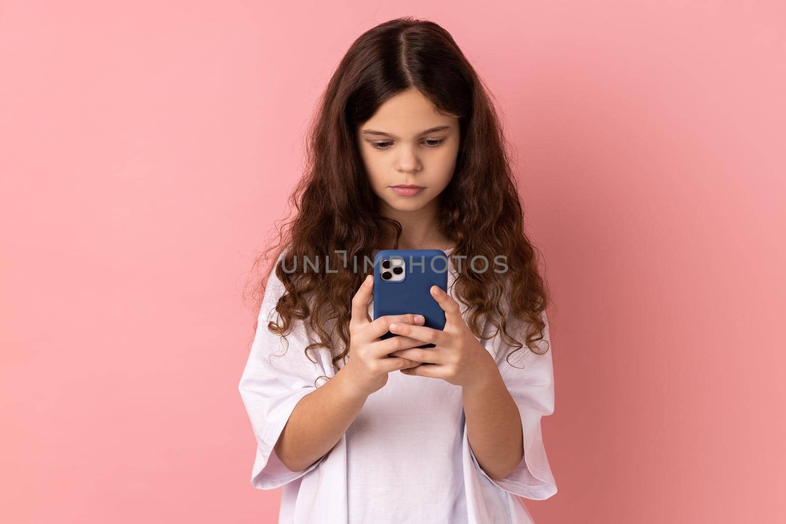 Portrait of serious little girl wearing white T-shirt texting message in social media on cell phone, using mobile network services, chatting online. Indoor studio shot isolated on pink background.