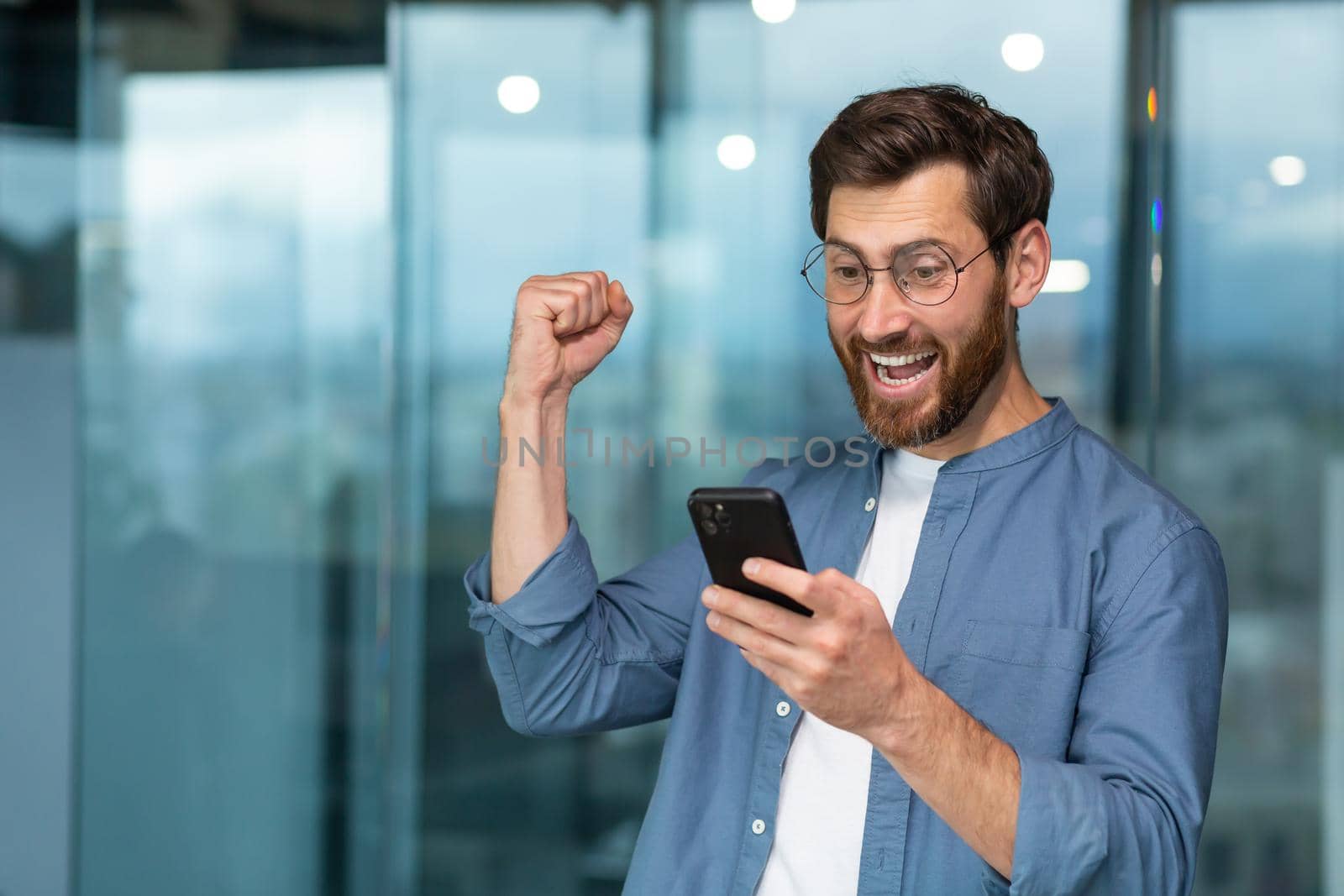 Successful businessman working in modern office building, man looking at smartphone and reading news celebrating happy news victory businessman in shirt and glasses holding hand up triumph gesture by voronaman