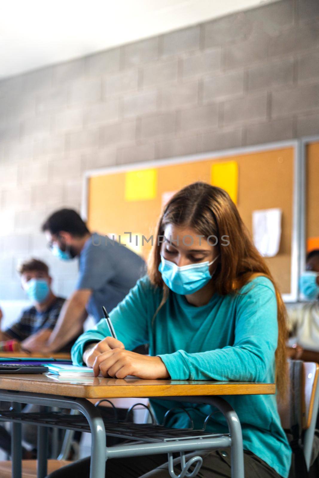 High school female student wearing face mask in class.doing homework. Teacher helping male teen student in background. Vertical image. Education concept. Healthcare concept.