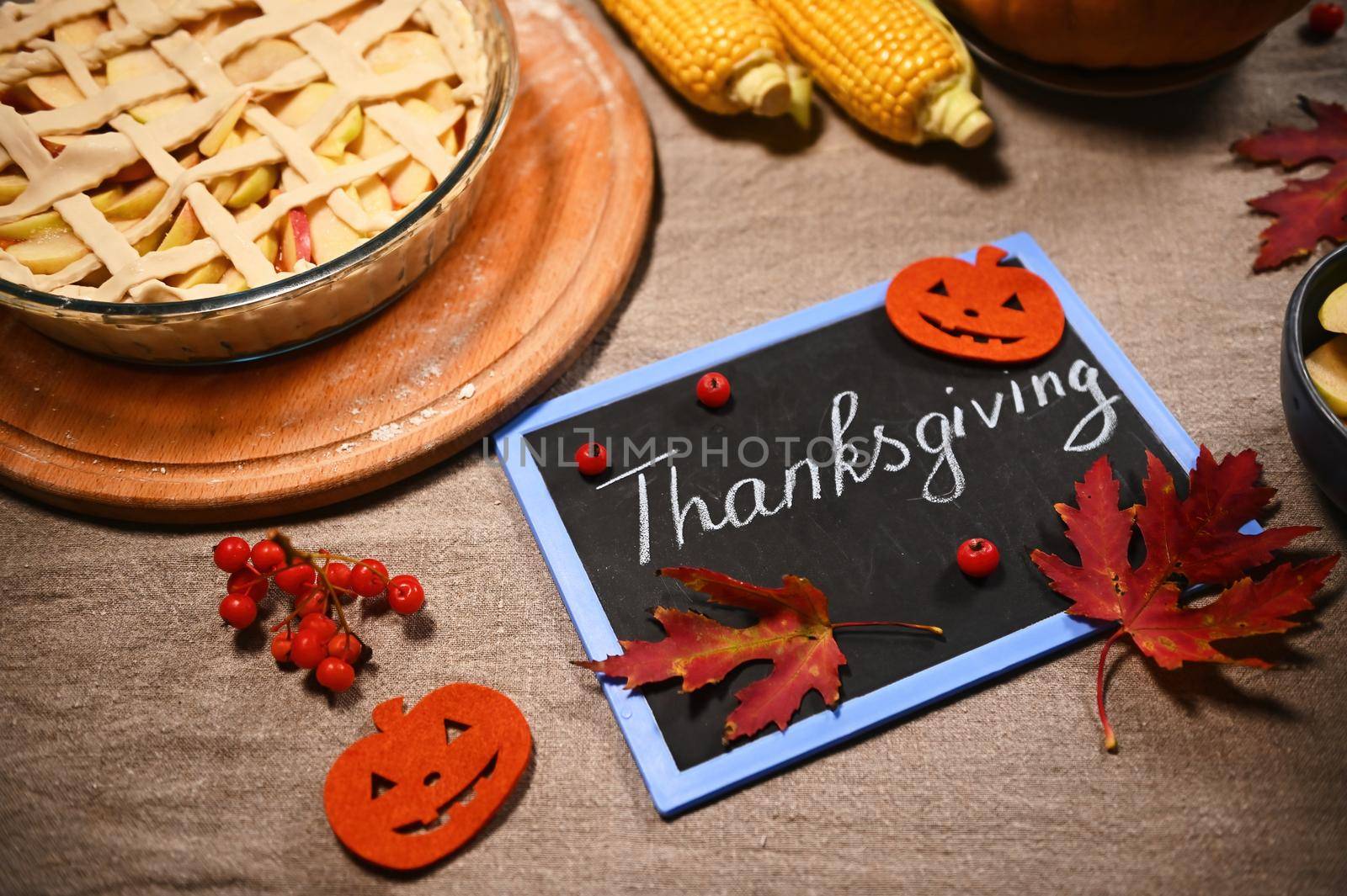 Blackboard with chalk lettering Thanksgiving Day, next to homemade festive pie and autumnal harvest of corn and pumpkin, on a table with linen tablecloth, dry fallen maple leaves and viburnum berries