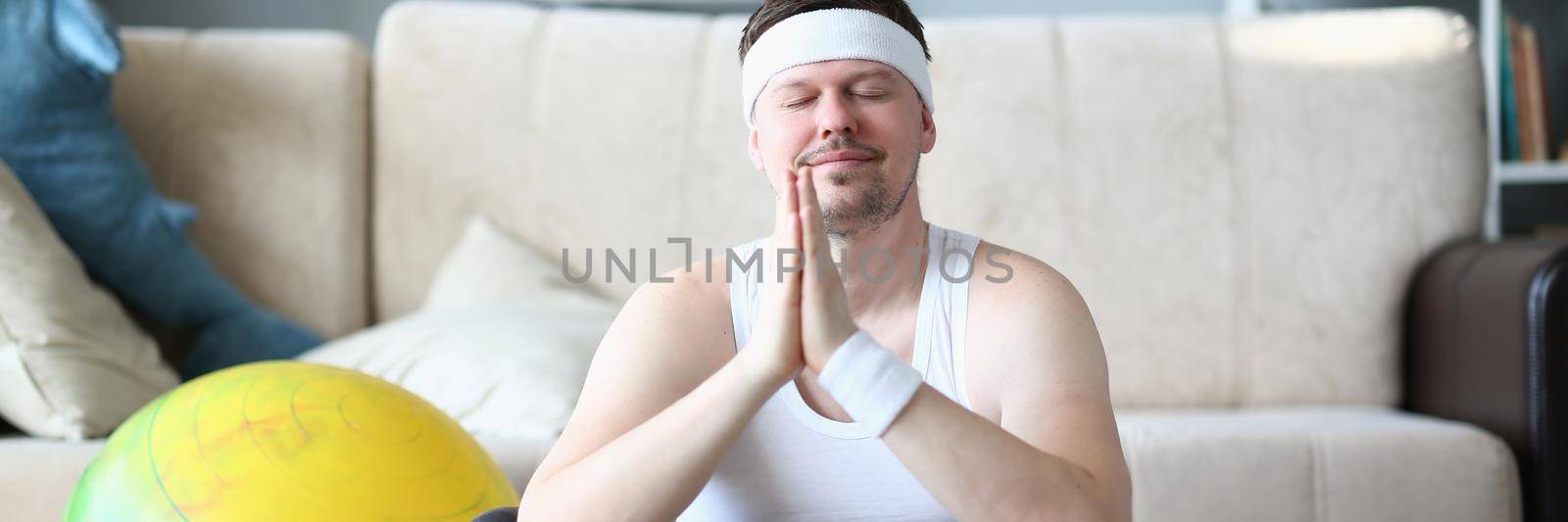 Man with praying hands and closed eyes meditates in lotus position at home. Exercises for relaxation and relieving emotional and muscle tension