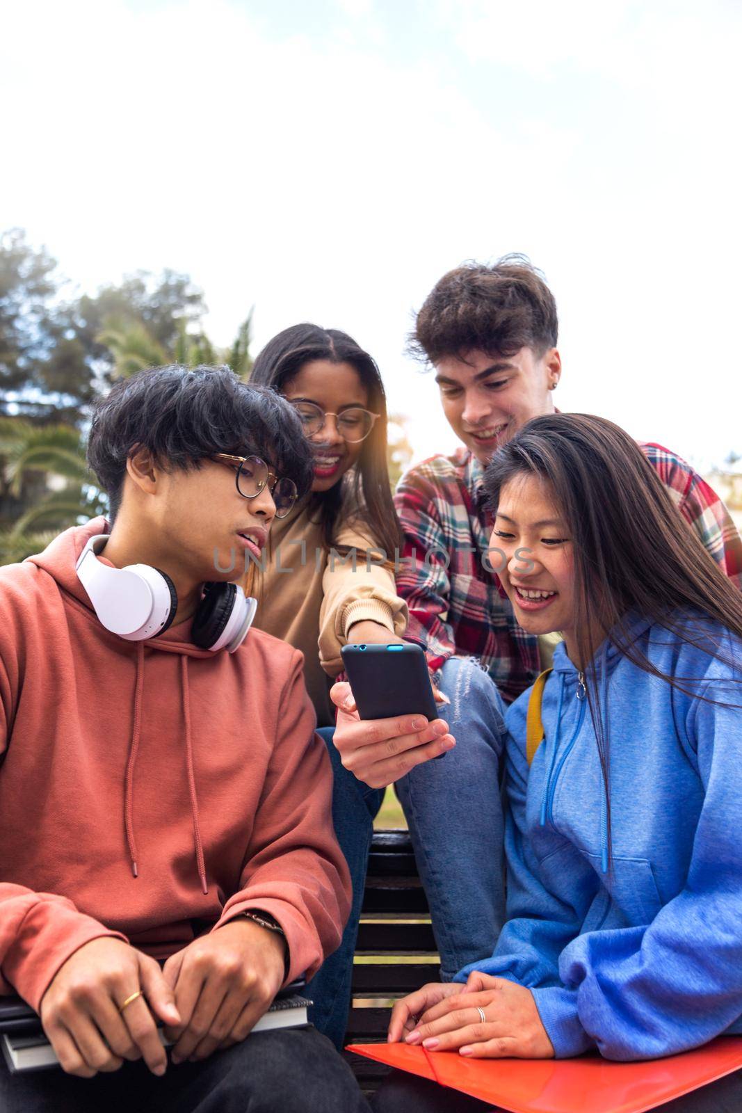 Group of multiracial happy young college student friends look at mobile phone. Teenagers using smartphone together outdoor. Vertical image. Youth lifestyle and social media addiction concept.
