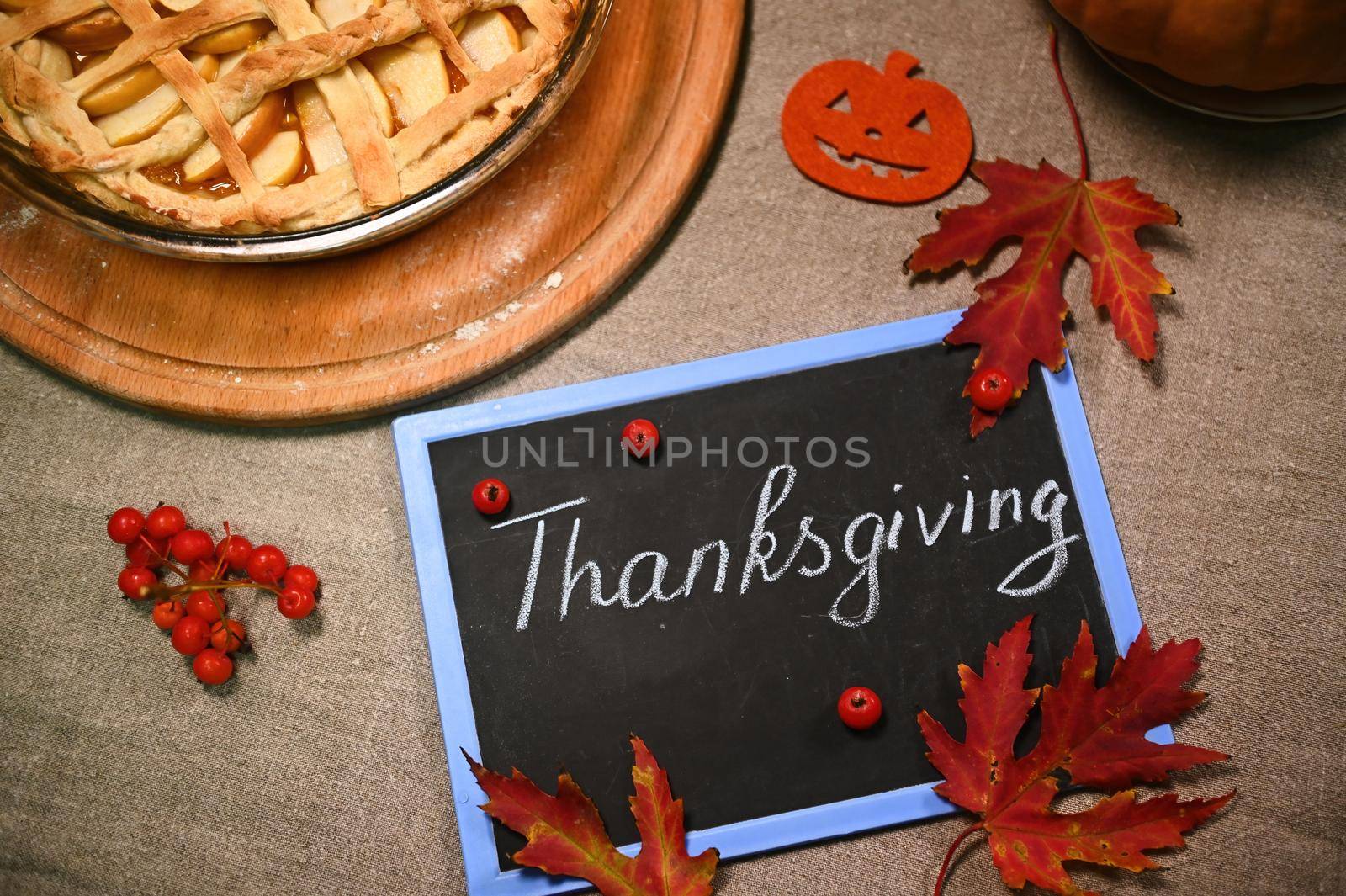 Selective focus. Dry fallen red autumn maple leaves and viburnum berries on a blackboard with chalk lettering Thanksgiving Day, on a linen tablecloth, next to a festive pumpkin pie with crust lattice