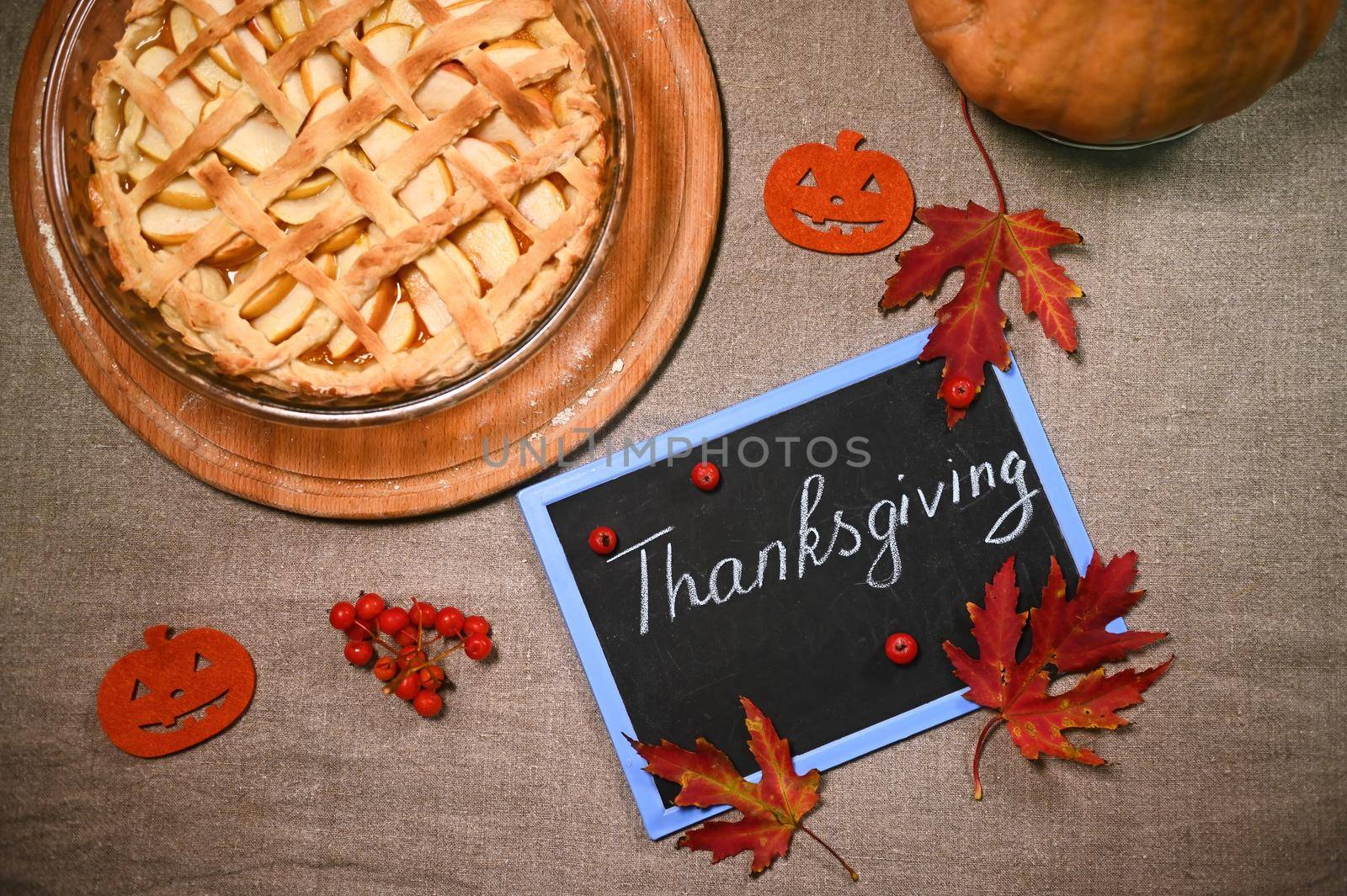 View from above. Dry fallen red autumn maple leaves and viburnum berries on a blackboard with chalk lettering Thanksgiving Day, on a linen tablecloth, next to a festive pumpkin pie with crust lattice