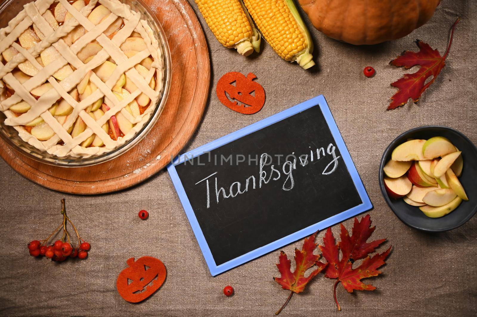 Top view of chalk board with chalk lettering Thanksgiving Day, next to a festive pie and a harvested autumn crop of corn and pumpkin lying on table with linen tablecloth and dry fallen maple leaves