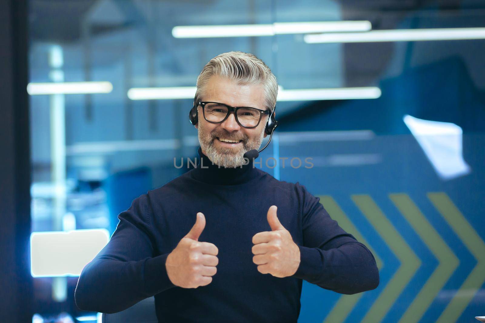 Motivational online training. A young handsome gray-haired male psychologist in headphones with a microphone conducts an online meeting, a webinar on camera. Super points with his fingers, smiles.