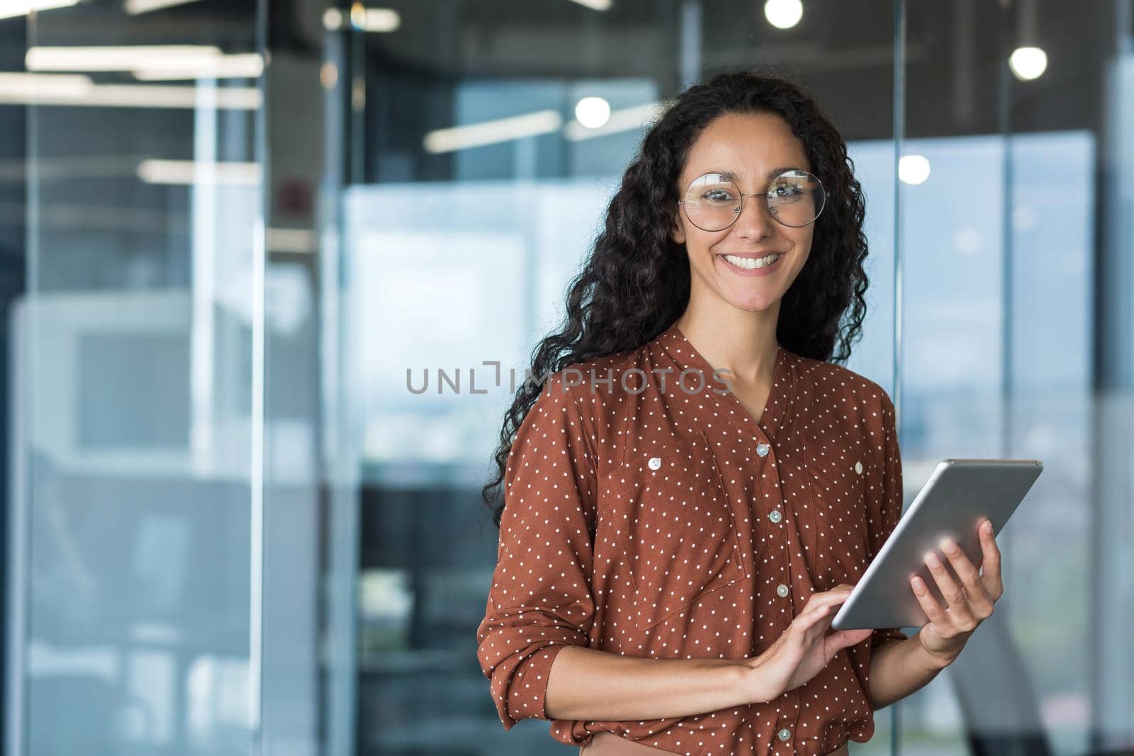 Portrait of a young beautiful Latin American woman journalist, writer. He is standing in the office, holding a tablet in his hands, typing information, looking at the camera, smiling.