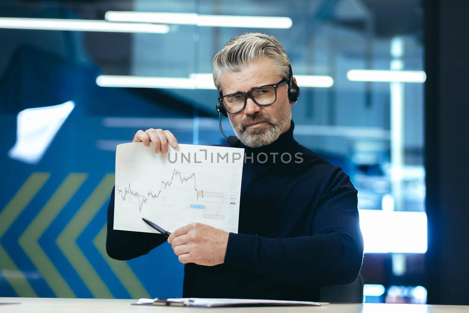 A serious businessman in headphones with a microphone conducts an online business meeting, a web conference, sits in the office, holds charts, a project in his hands and points to the camera. by voronaman