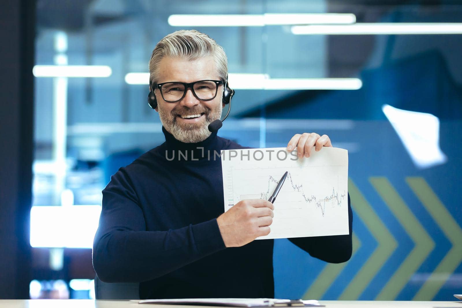 Portrait of a man in headphones with a microphone on his head. Sitting in the office, holding graphics, a project in his hands by voronaman