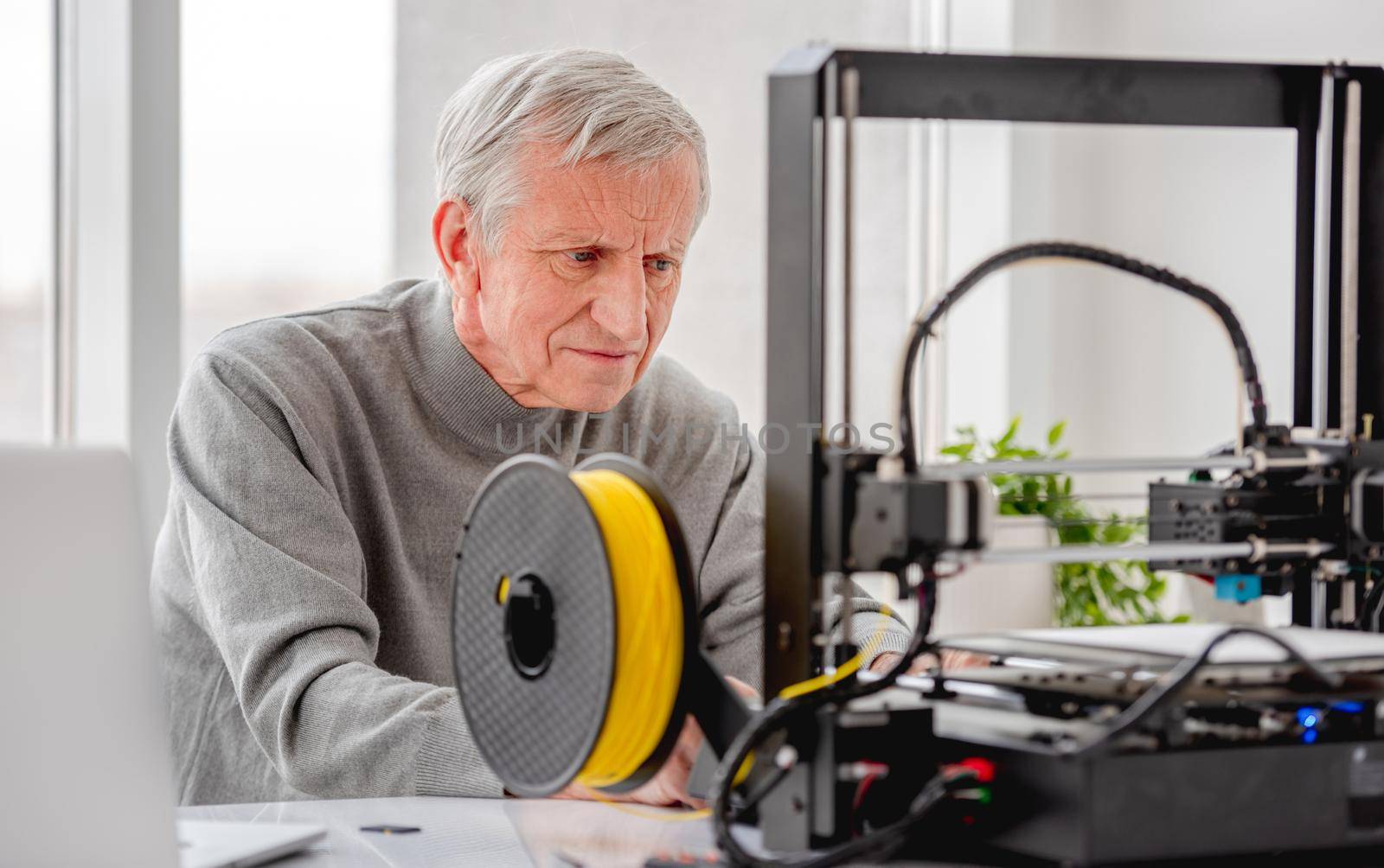 Adult man sitting at labaratory with daylight and looking at modern 3D printer