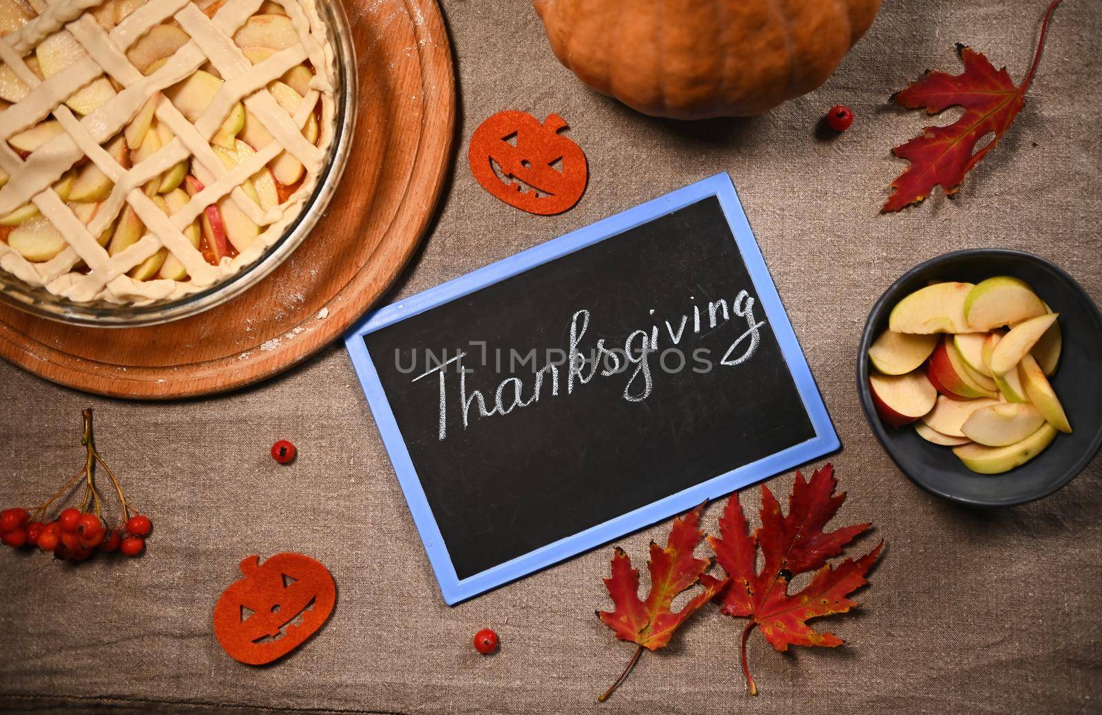 Top view of a chalkboard with Thanksgiving lettering next to dry autumn maple leaves, viburnum berries, sliced apple in blue ceramic bowl and homemade holiday pumpkin pie with crispy crust. Still life