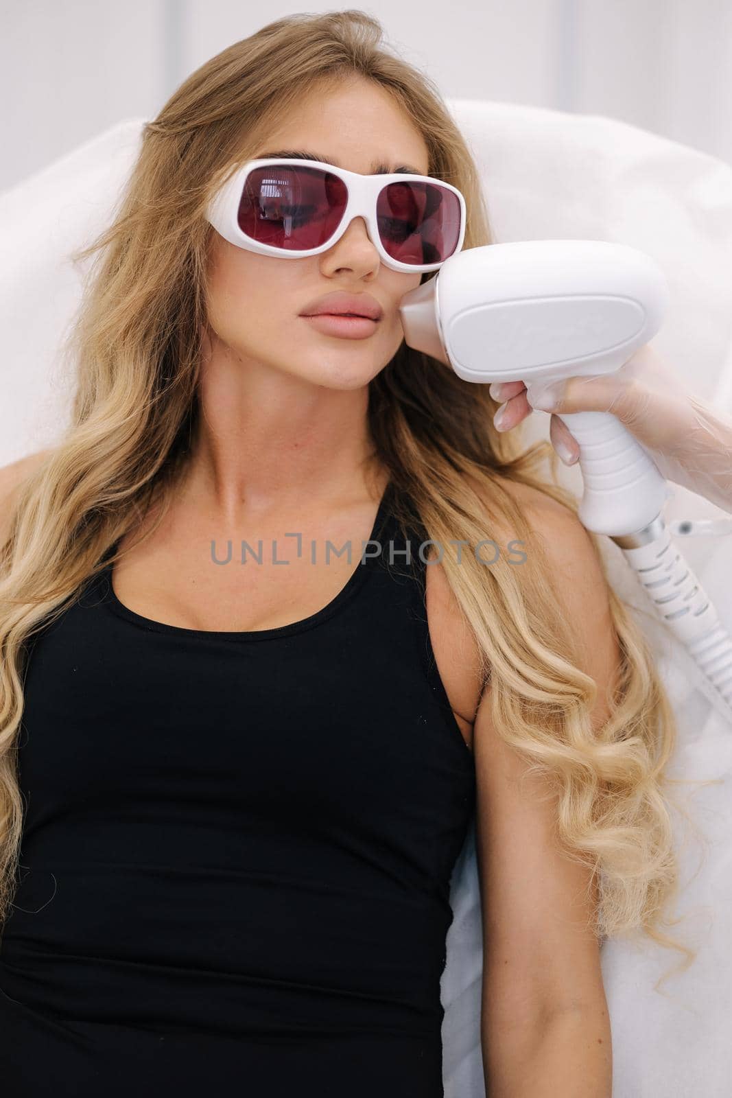 Beautician doing epilation on the face of a beautiful woman in beauty center. Female receiving laser light hair removal treatment for hairless smooth skin at cosmetology salon. Model in protective glasses. Close-up by Gritsiv