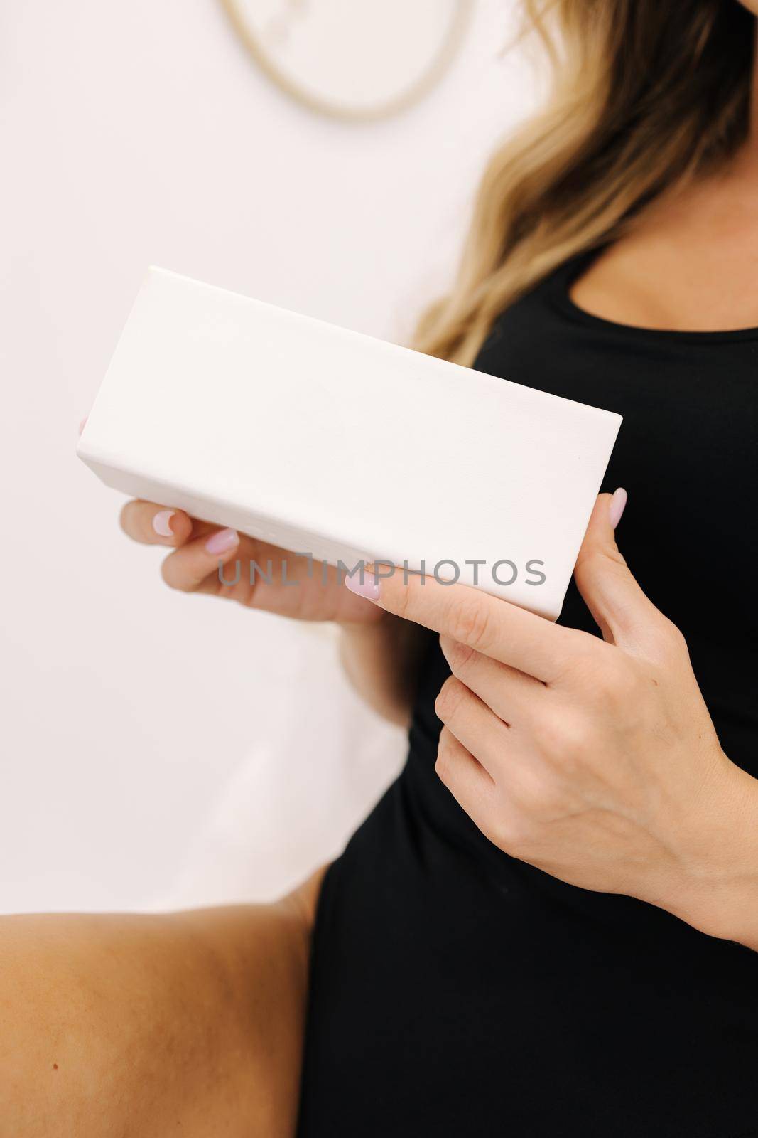 Beautiful female model in cosmetology studio hold white box in a hand. Sexy woman in black bodysuit. Advertising of beauty product.