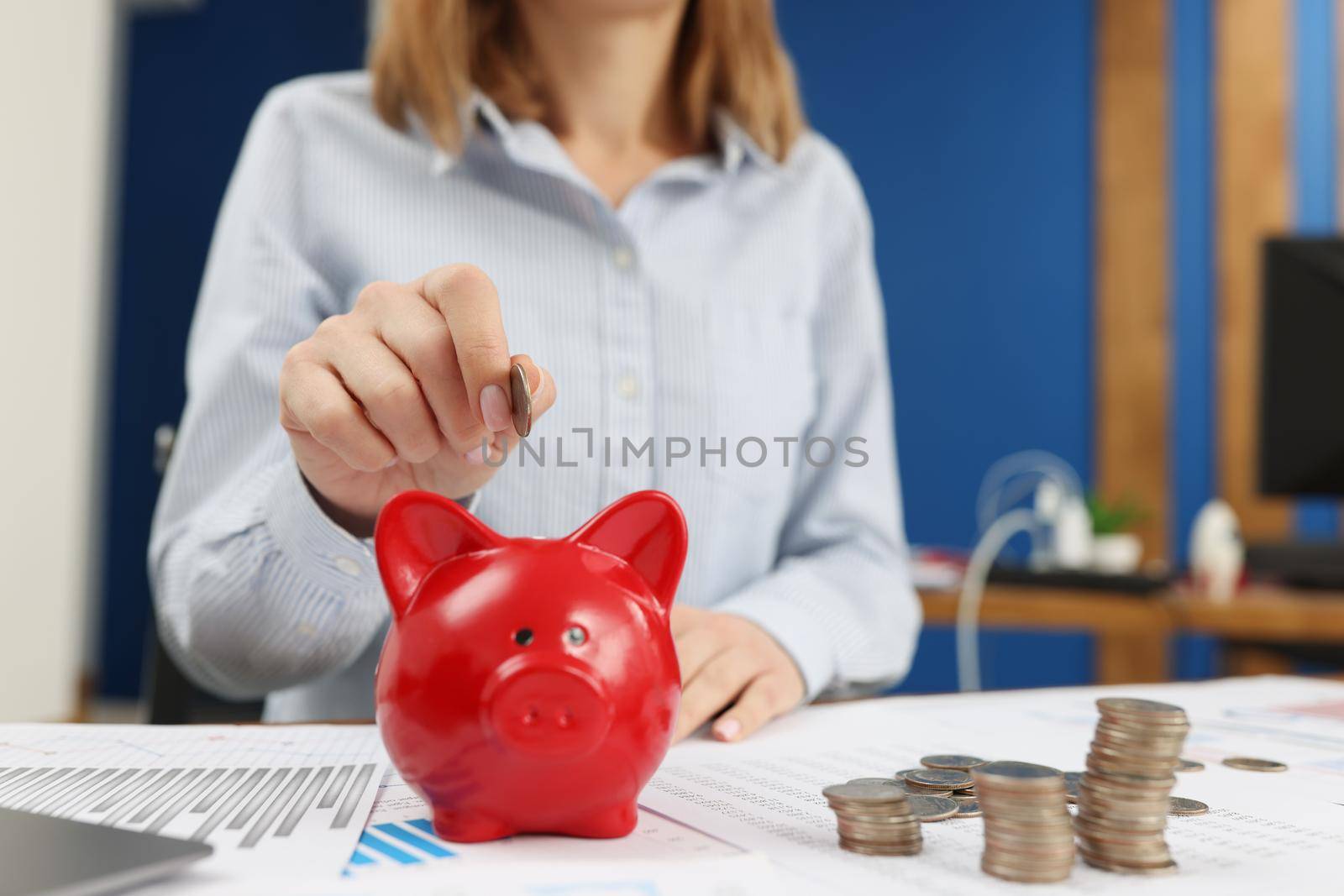 A woman in the office puts a coin in a red piggy bank, close-up. Capitalization of income, saving money, investments