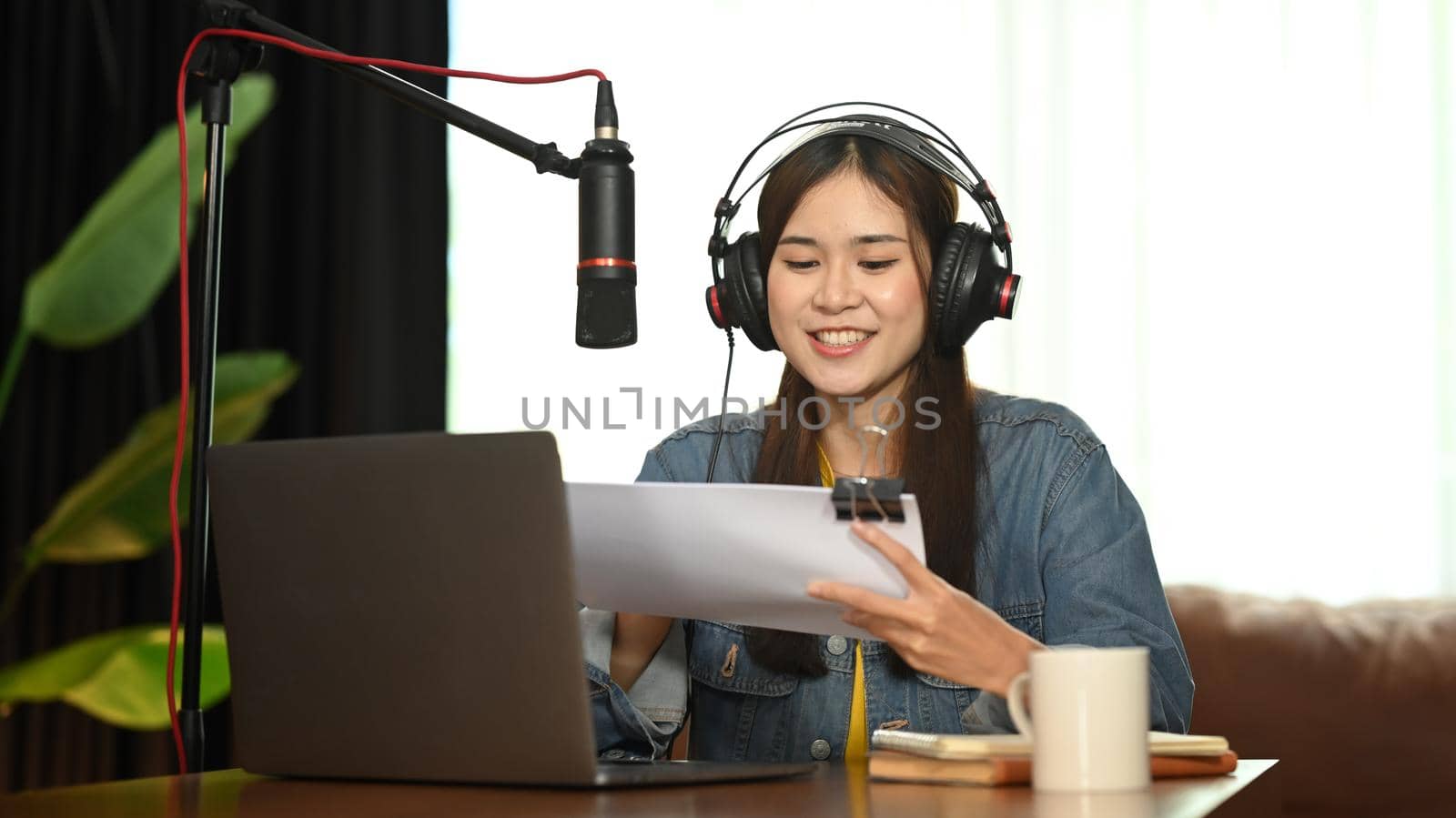 Smiling asian woman podcasters using laptop and condenser microphone to recording podcast in small home studio.