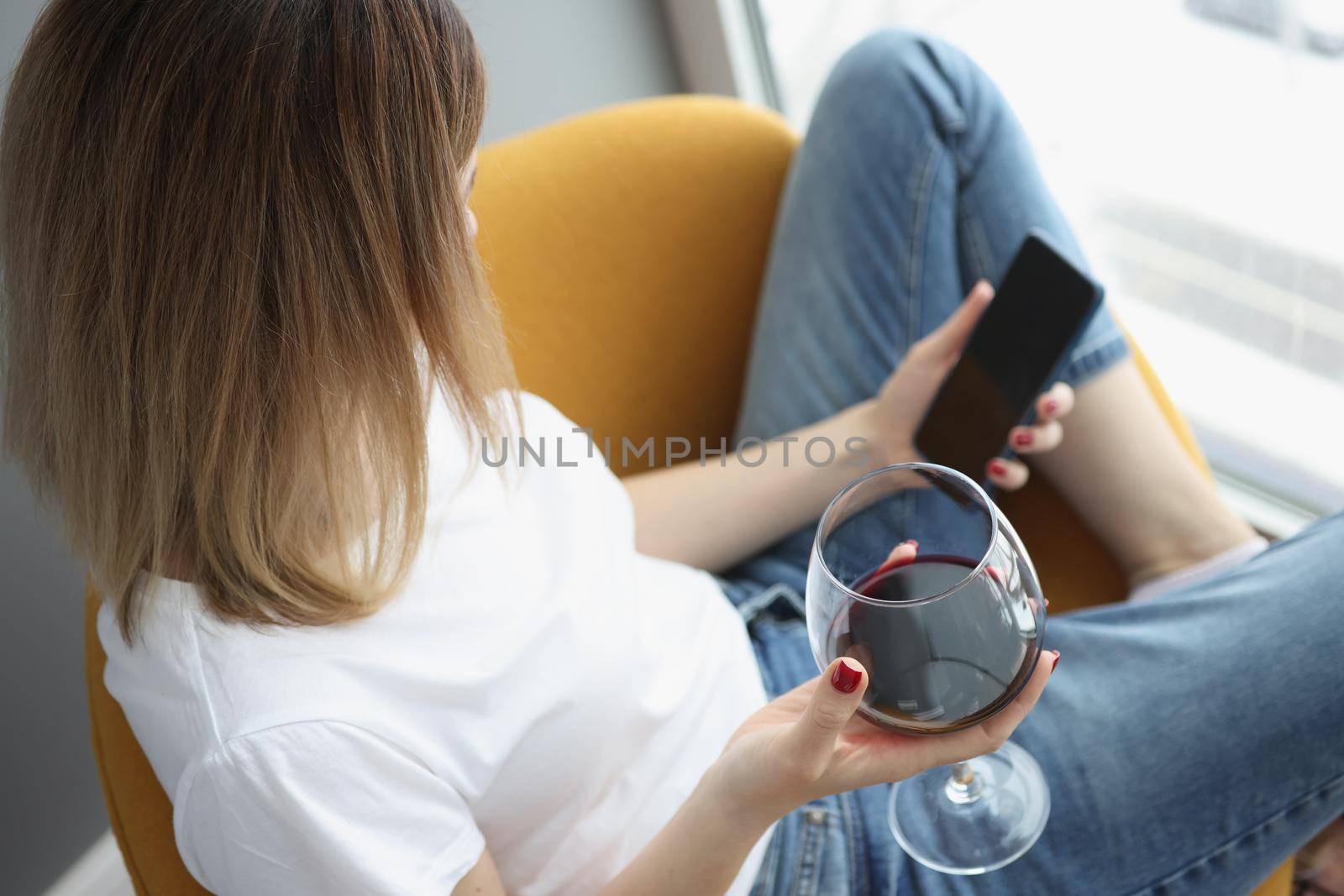 A woman at the window holds a glass of red wine and a smartphone, top view, close-up. Loneliness, stress relief, self-isolation