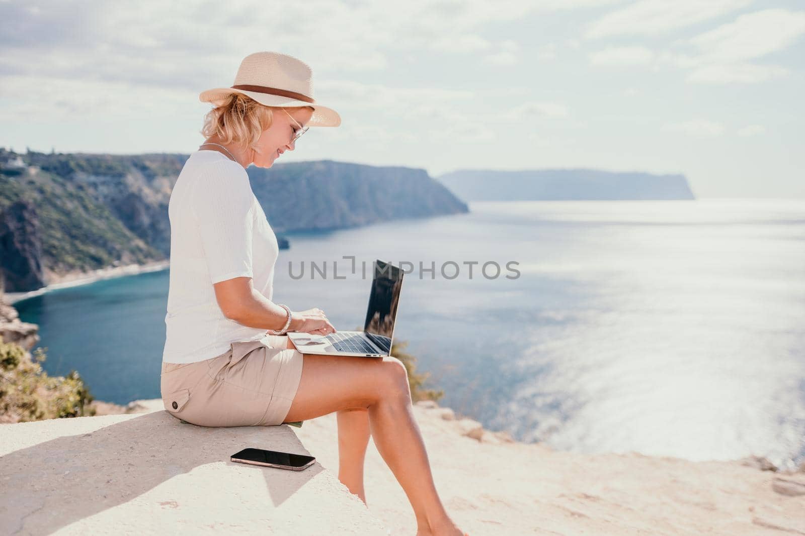 Successful business woman in yellow hat working on laptop by the sea. Pretty lady typing on computer at summer day outdoors. Freelance, travel and holidays concept.