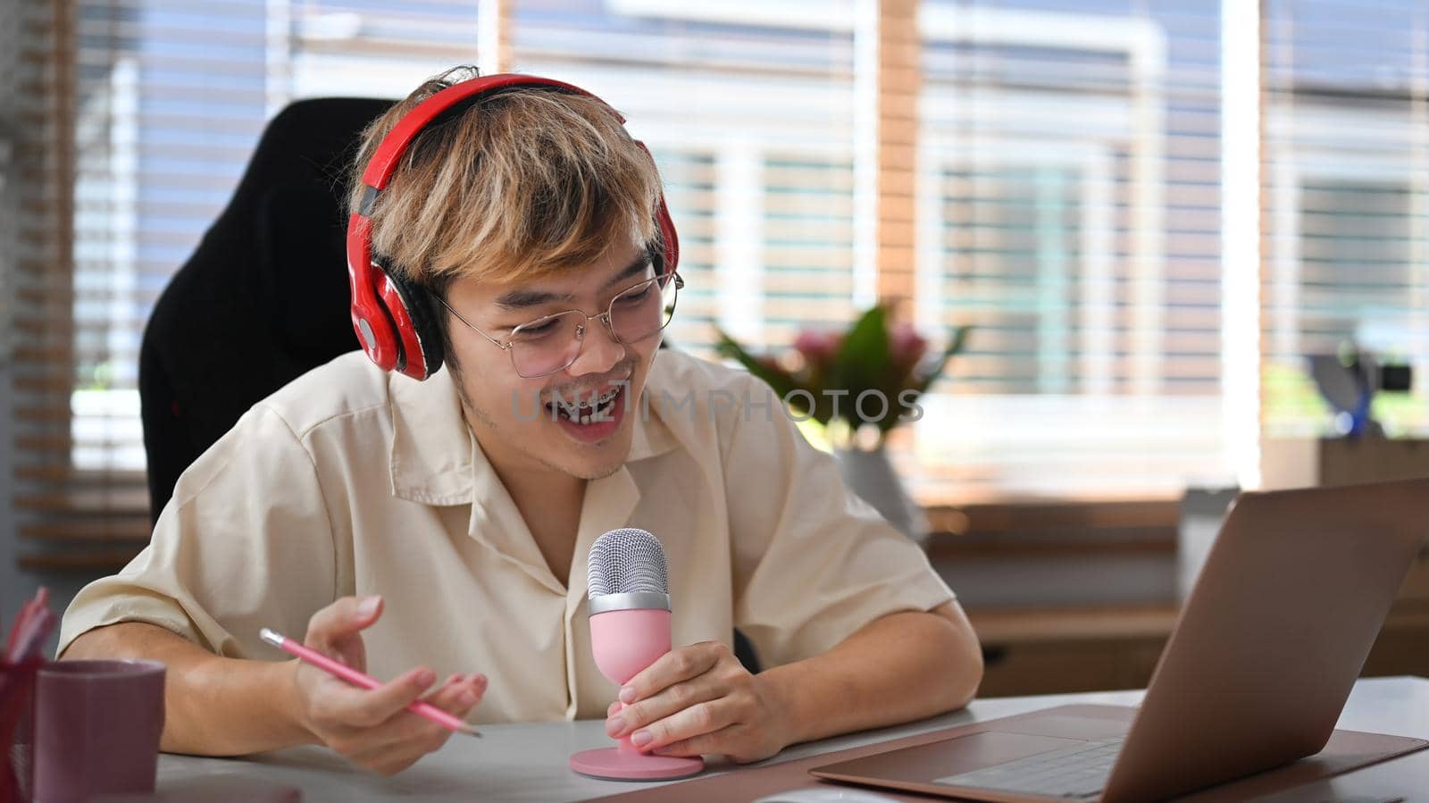 Cheerful young asian man using laptop and microphone streaming audio podcast at home studio.