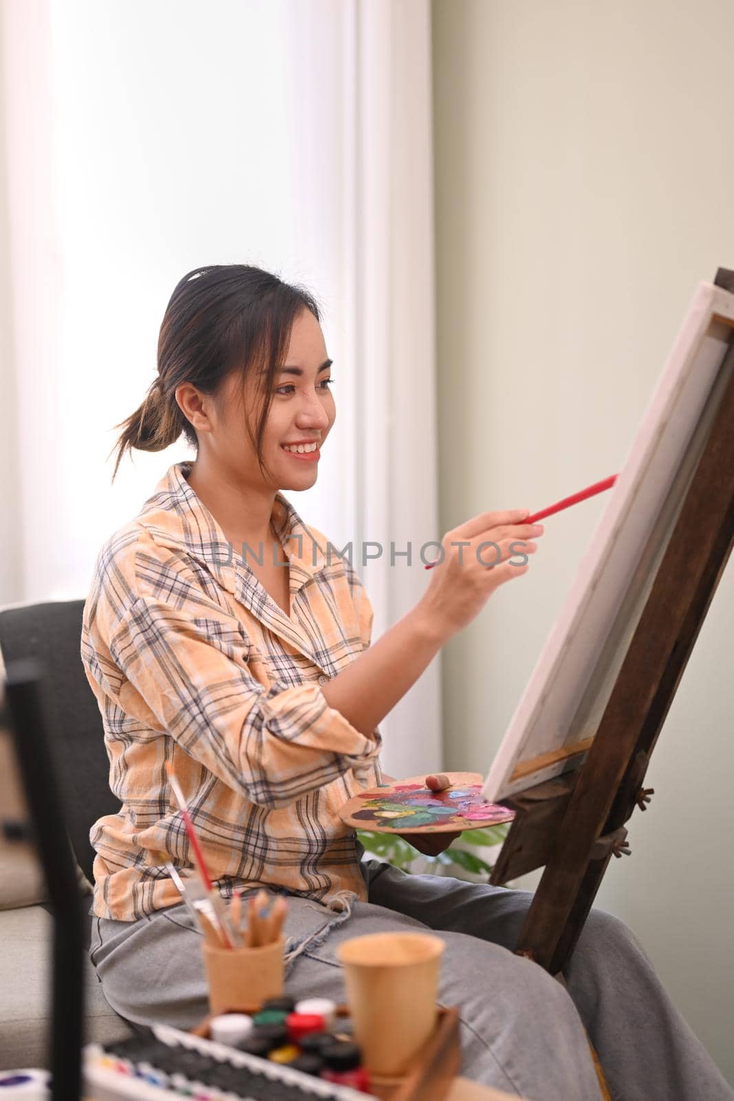 Satisfied millennial woman painting picture on canvas with watercolor in bright home studio. Leisure activity and art concept.