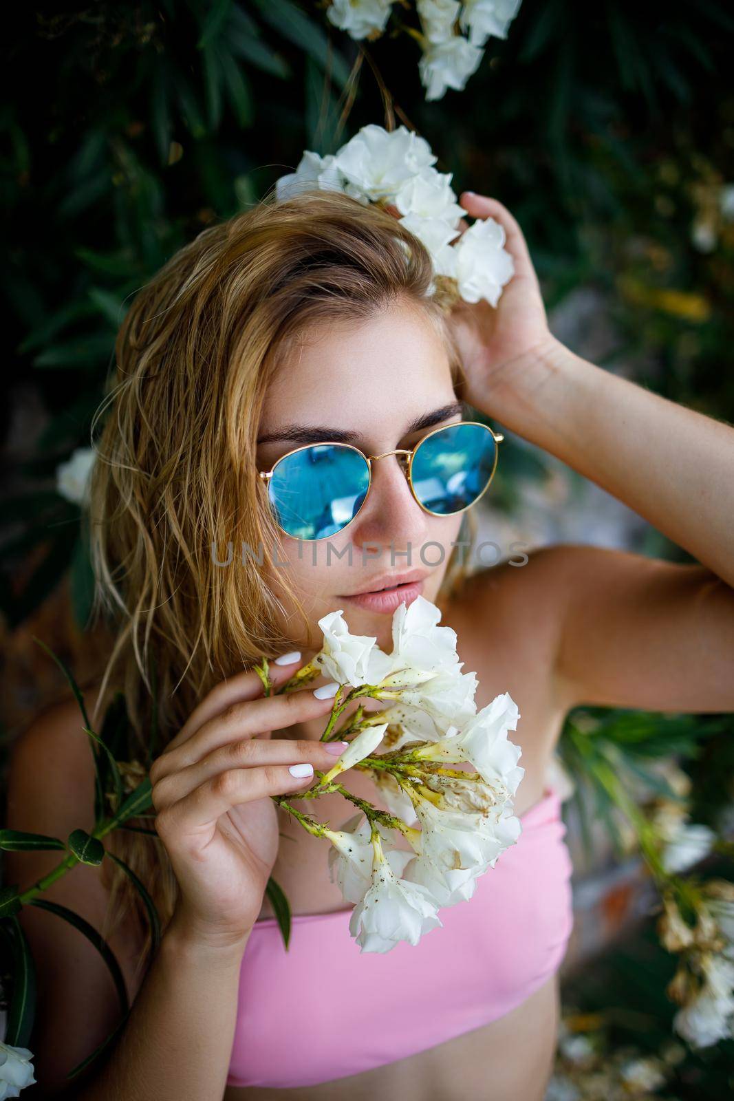 Attractive young woman in a pink swimsuit, sunglasses on her face stands by a plant with white flowers by Dmitrytph