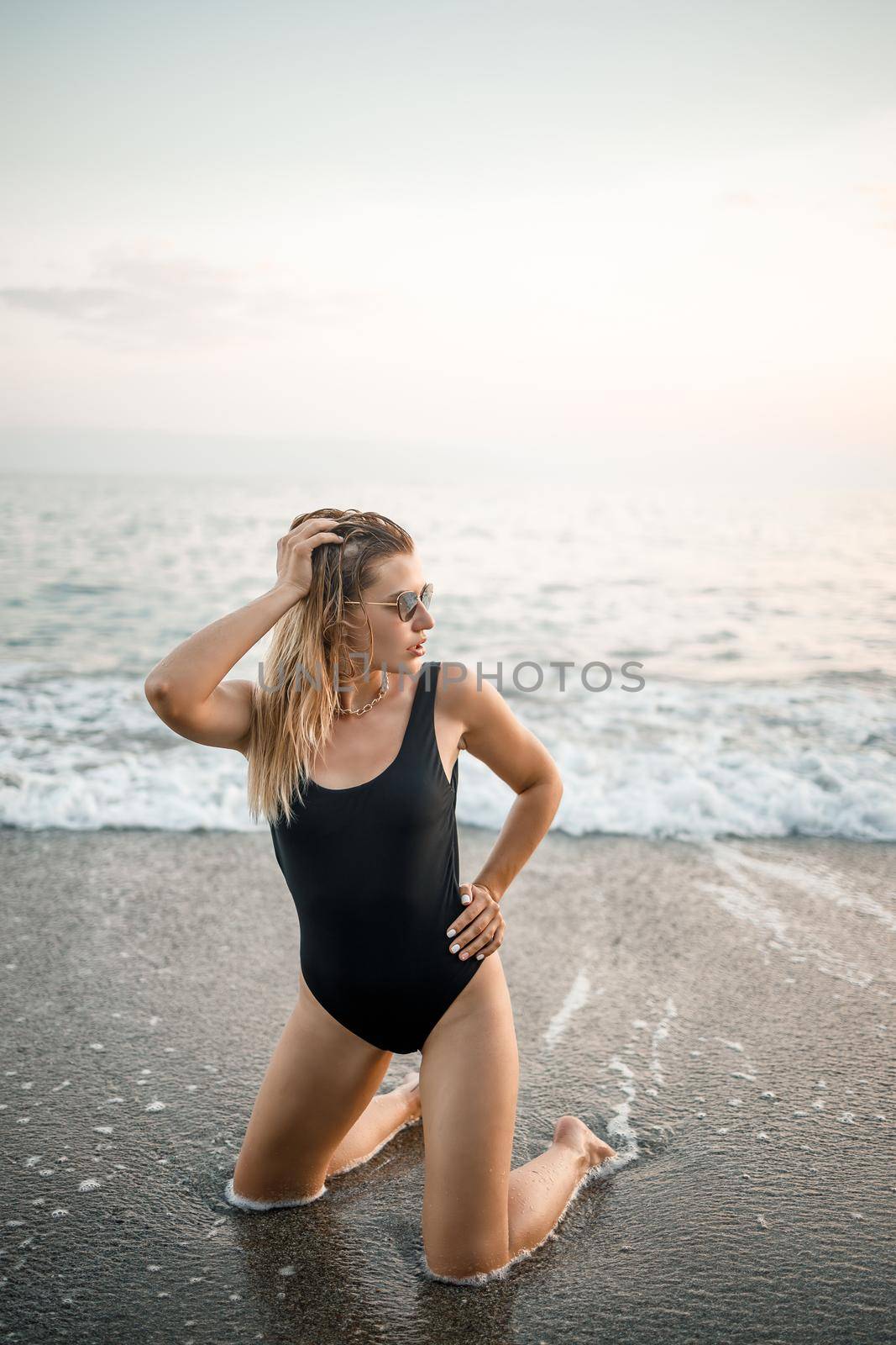 Attractive young woman in sunglasses kneels in the sea at sunset light in a black swimsuit. Selective focus by Dmitrytph