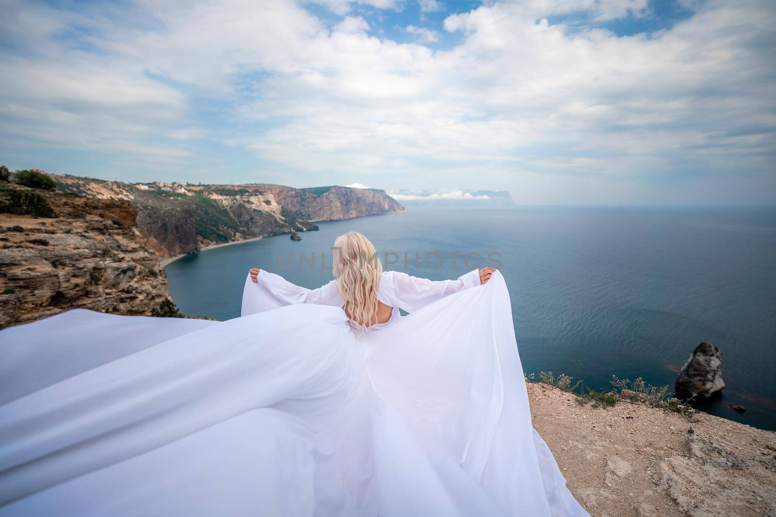 Blonde with long hair on a sunny seashore in a white flowing dress, rear view, silk fabric waving in the wind. Against the backdrop of the blue sky and mountains on the seashore. by Matiunina