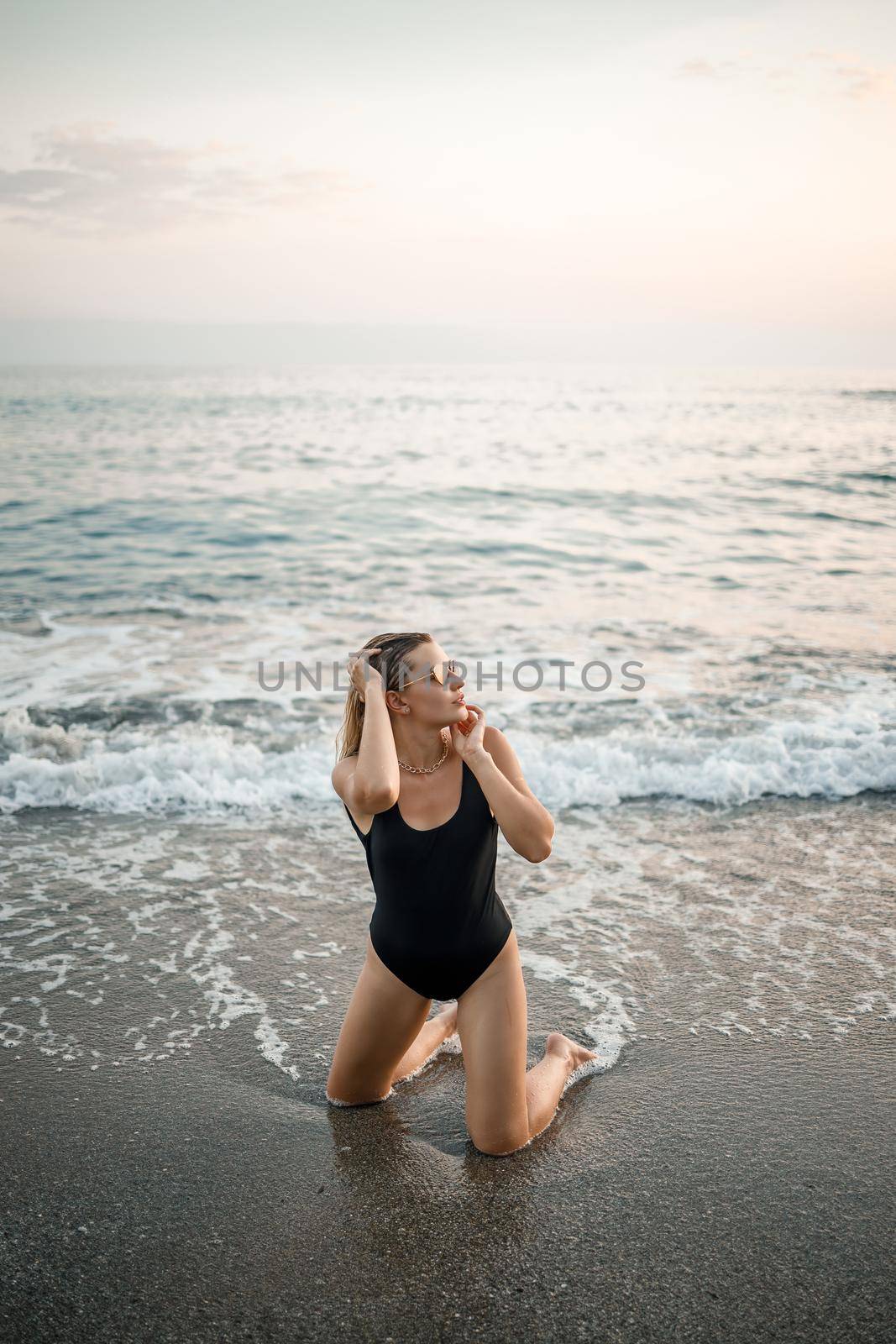 Attractive young woman in sunglasses kneels in the sea at sunset light in a black swimsuit. Selective focus by Dmitrytph