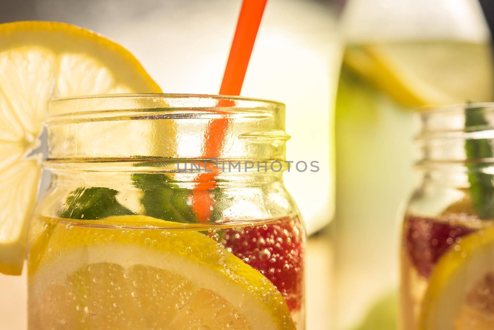close-up of a glass jar lit by sunlight with refreshing cold lemonade water, lemon slices, red berries, mint leaves and drink cane. Summer citrus soda background