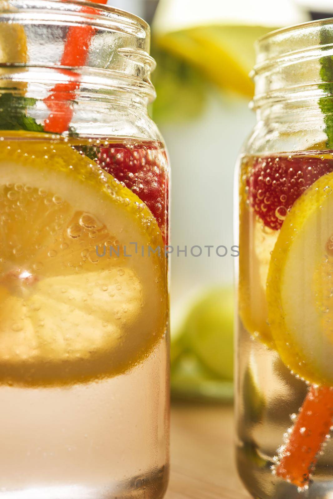 two glass jars and a bottle lit by sunlight with refreshing cold lemonade water, lemon slices, red berries, mint leaves and drink canes on a wooden table. Summer citrus soda background, vertical photo