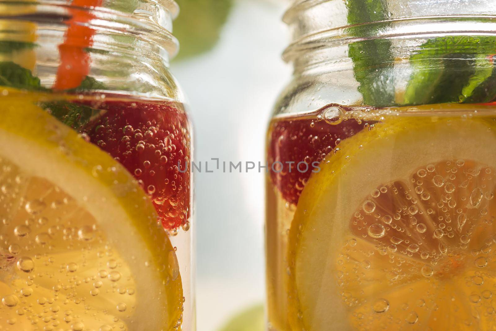 close-up of two glass jars illuminated by sunlight with refreshing cold lemonade water, lemon slices, red berries, mint leaves and drinking cane. Summer citrus soda background