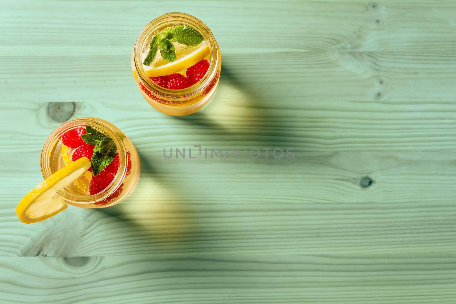 overhead shot of two glass jars with cold water, lemon slices, red berries and mint on one side of a green wooden table illuminated by sunlight. Summer refreshment background with citrus, copy space