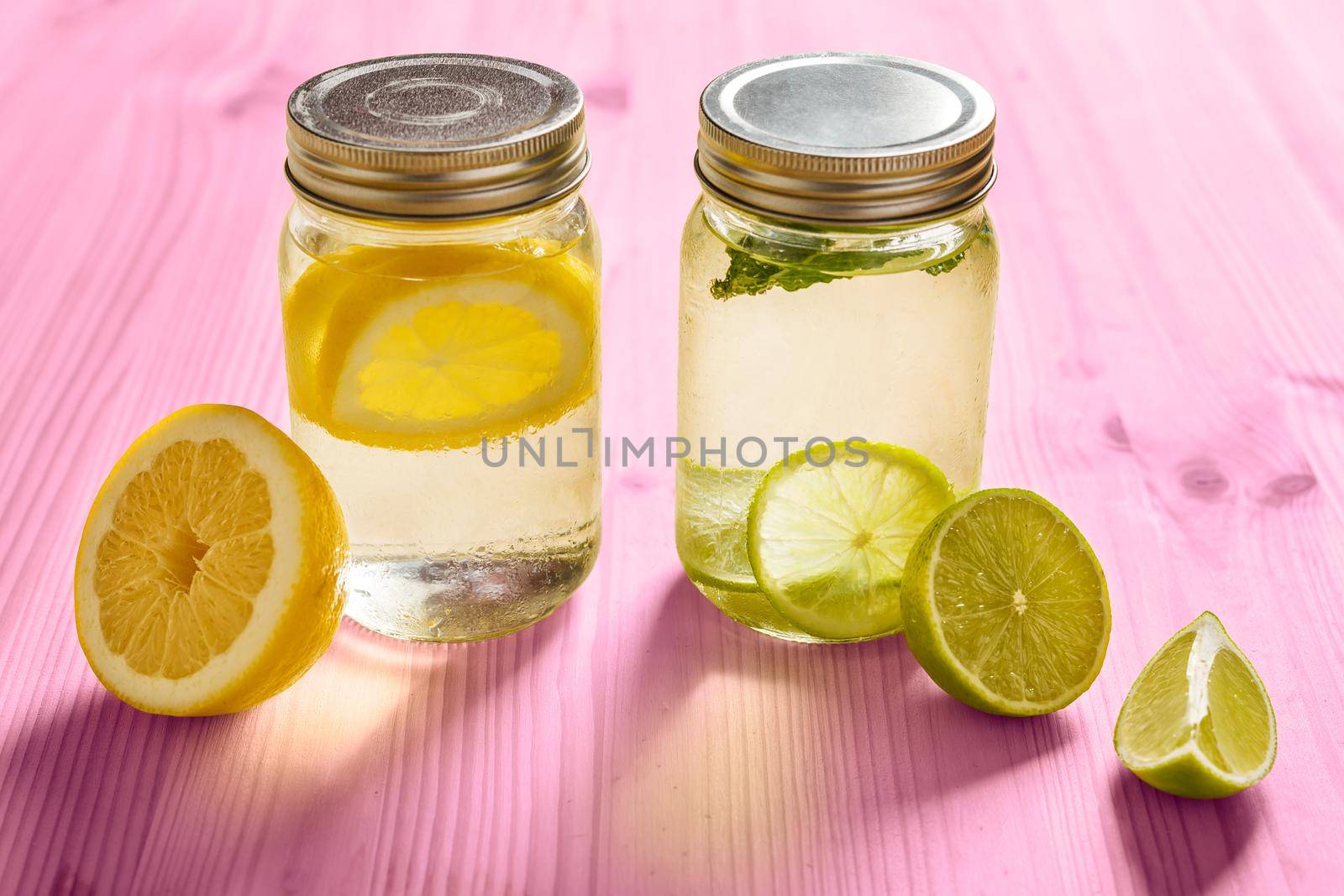 two glass jars with lid, one with cold water and slices of lemon and another with slices of lime and mint, are illuminated by sunlight and on a pink wooden table with some pieces of citrus fruits