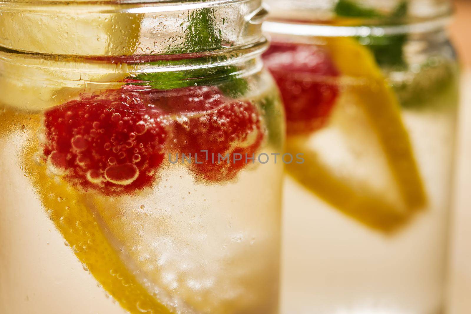 close-up of a glass jar with cold water, slices of lemon, mint and red berries, illuminated by sunlight on a wooden table, in the background you can see out of focus another glass equal