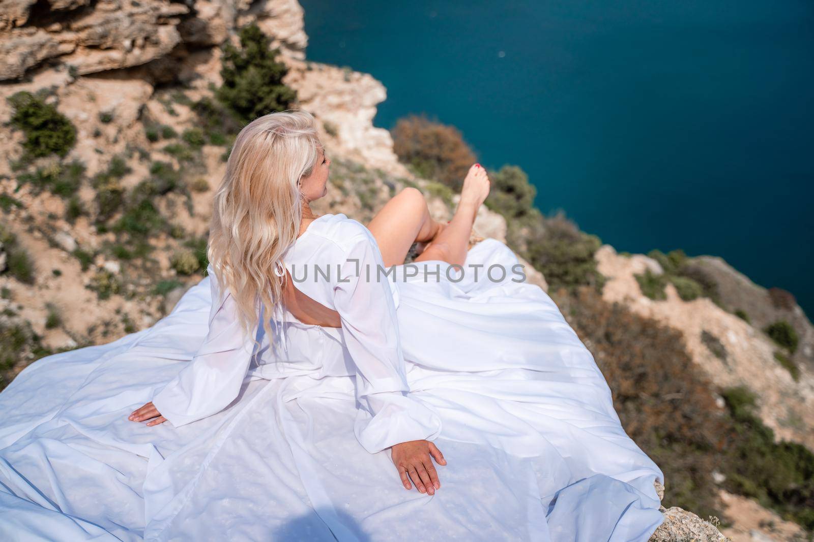 The blonde sits on a rock above the sea. With long hair on a sunny seashore in a white dress, rear view, silk fabric flutters in the wind. Against the backdrop of blue skies and mountains. by Matiunina