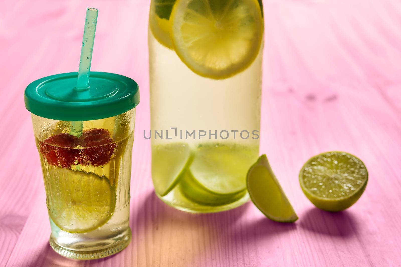 bottle and glass with lid and cane with cold water and slices of lime, lemon and red berries, illuminated by sunlight on a pink wooden table with some pieces of citrus, summer refreshments background